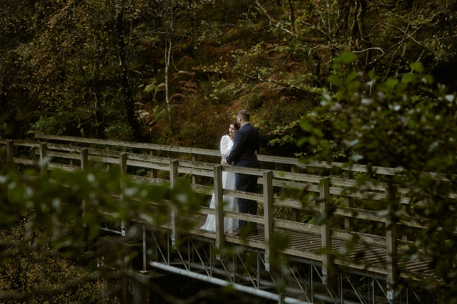 Couple standing on wooden bridge during Glencoe Lochan elopement surrounded by autumn forest
